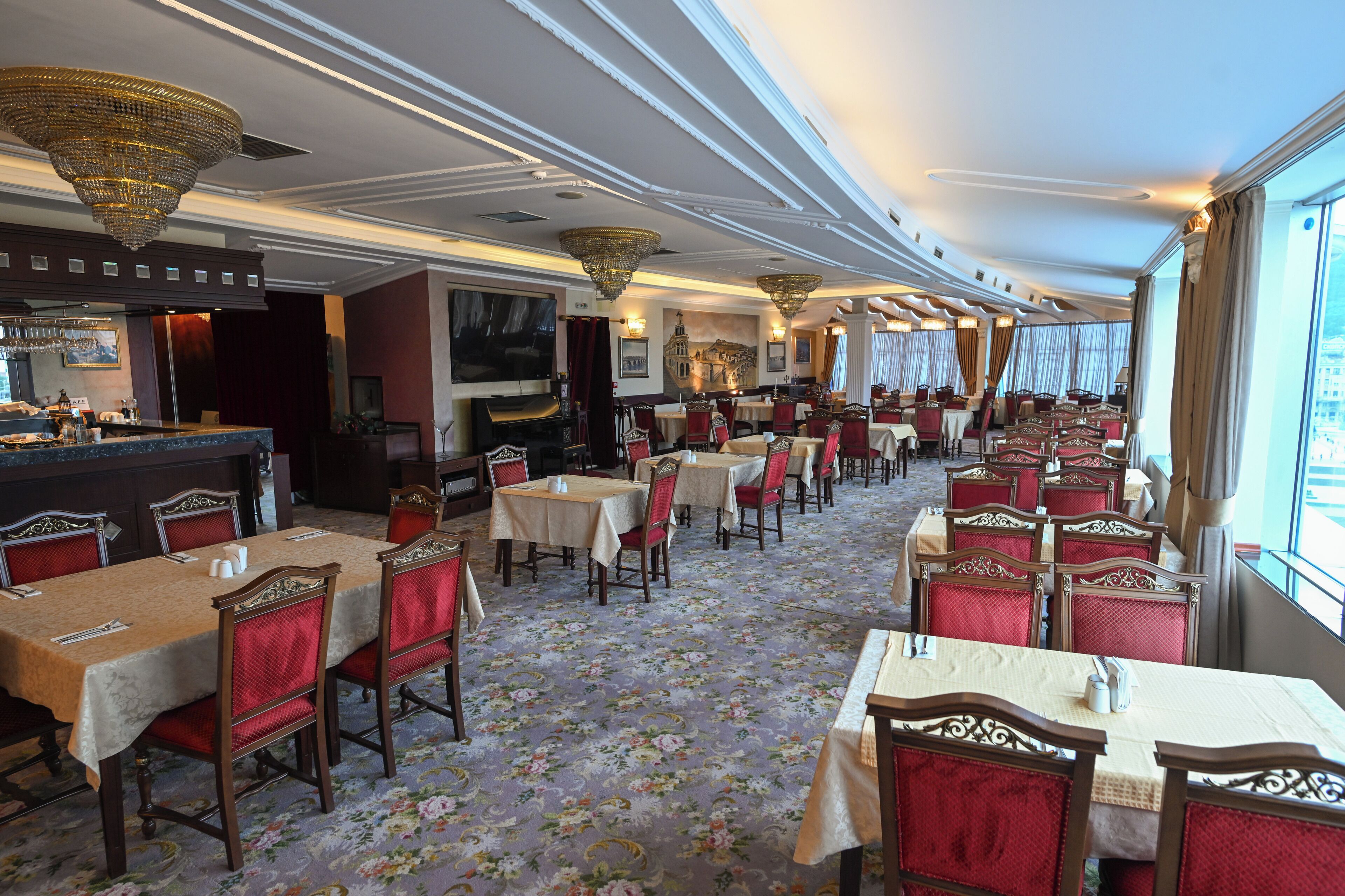 A spacious restaurant with multiple tables and chairs, a bar area, and large windows offering a view.