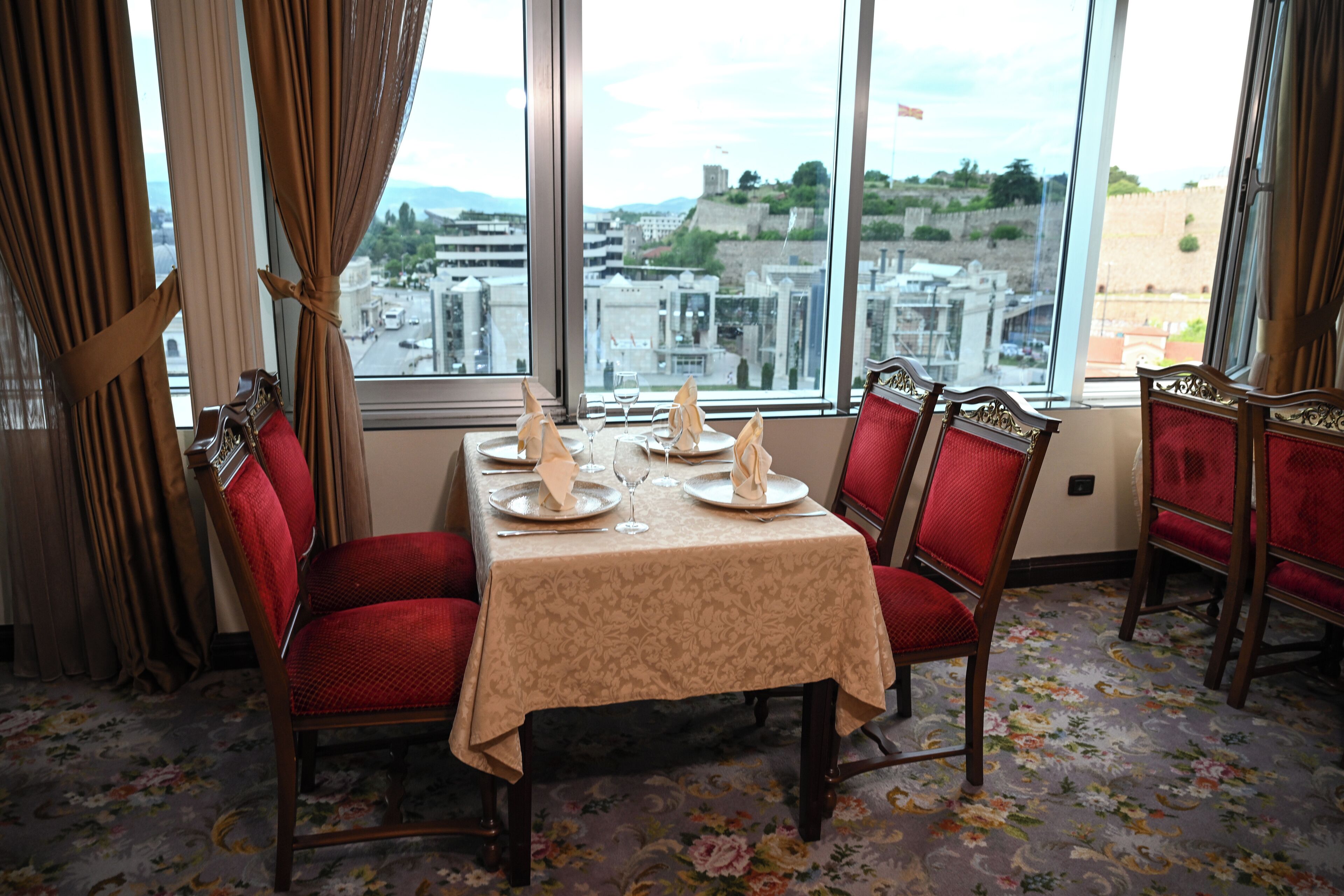 Une salle à manger avec une table dressée pour quatre personnes, des chaises à l’assise rembourrée de tissu rouge et une vue sur la ville grâce à de grandes fenêtres.