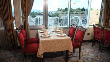 A dining room with a table set for four, red upholstered chairs, and a view of the city through large windows.