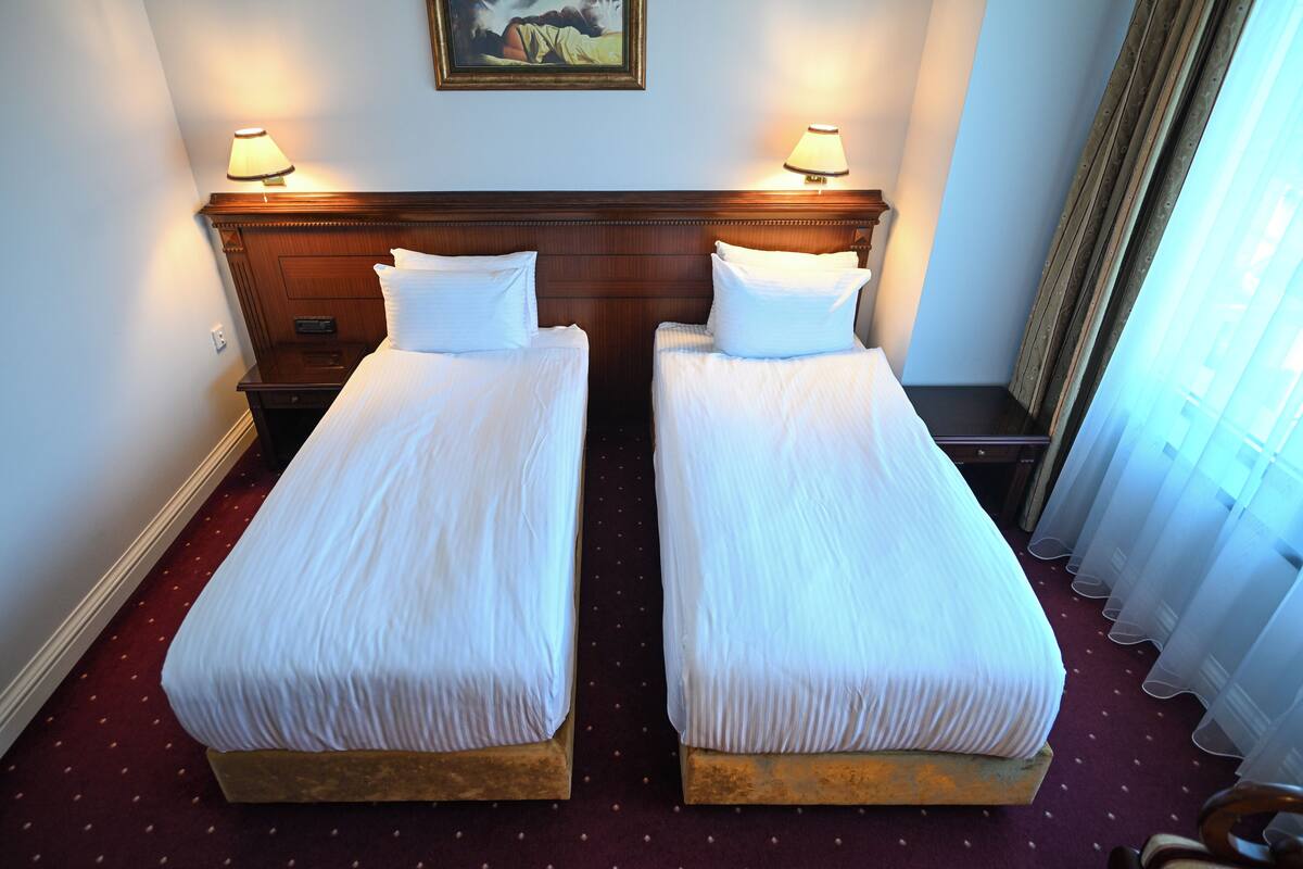 A hotel room with two single beds, wooden headboard, a nightstand, a lamp, and a painting on the wall.