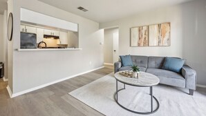 A modern living room with a grey sofa, a round coffee table, and a kitchen area visible through an open door.
