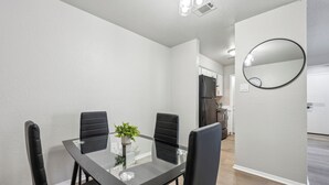A modern dining area with a glass table and black chairs, a small potted plant, and a kitchen in the background.