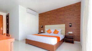 Une chambre d’hôtel moderne avec une tête de lit en bois, un grand lit avec une literie blanche et des touches de couleur orange, une table de chevet en bois et une petite étagère.