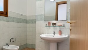 A bathroom with a white pedestal sink, a bidet, and a glass shelf with toiletries. - [Green Hotel] Rural hotel double rooms (Orosei)