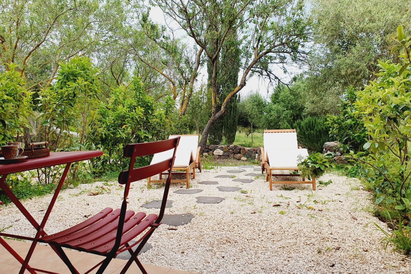An outdoor seating area with a red table and chairs, surrounded by greenery and a stone pathway.