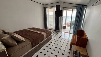 A bedroom with a bed, a desk, a chair, a fan, and a view of the sea through a window.