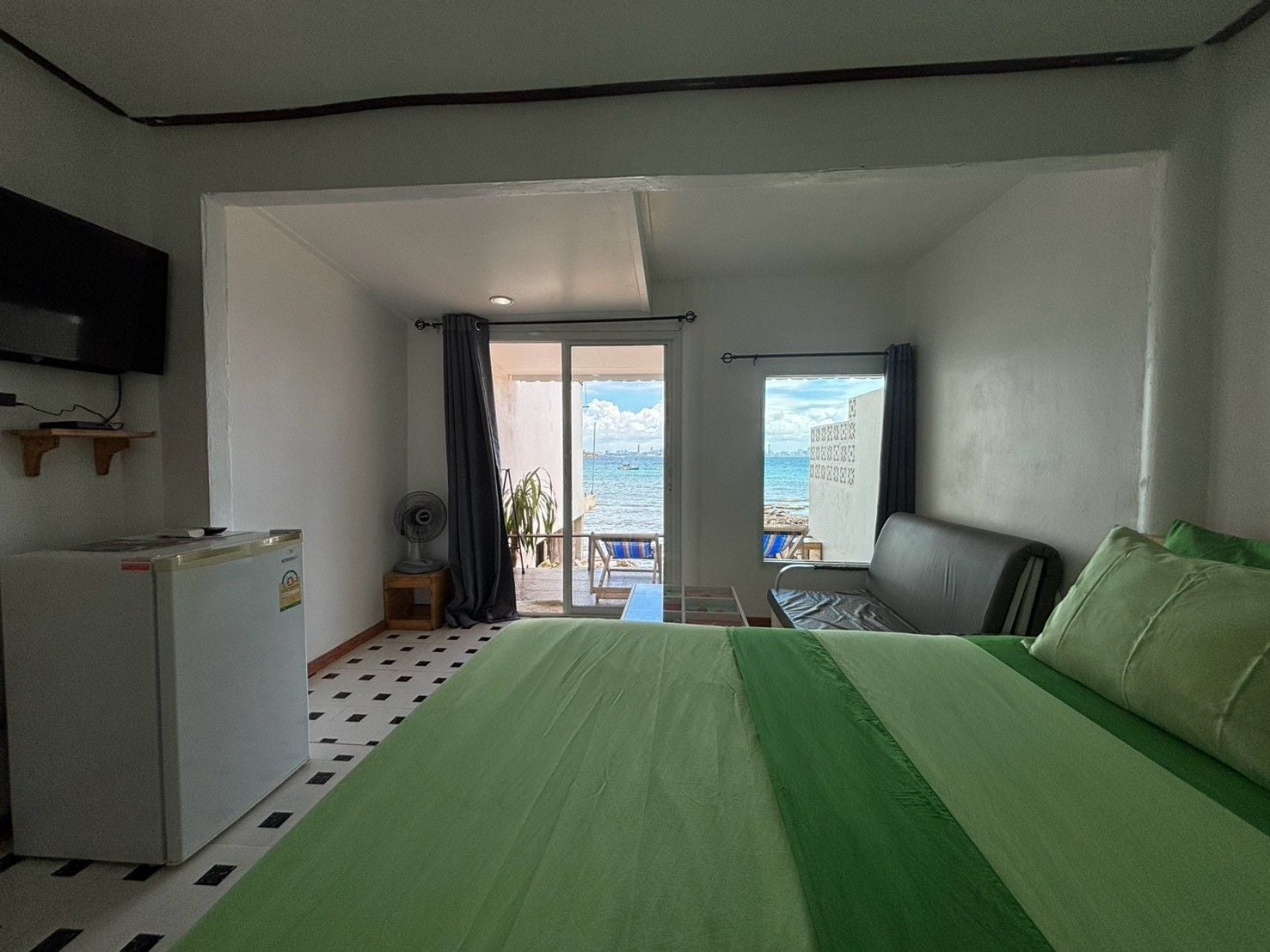 Een hotelkamer met een bed, een bureau, een stoel, een klein tafeltje, een ventilator en uitzicht op zee.