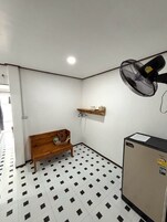 A small room with a wooden bench, a wall-mounted shelf, a standing fan, and a refrigerator.