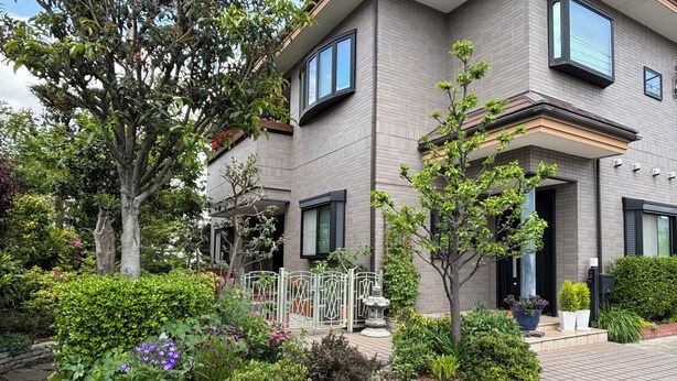 A modern two-story house with a brick exterior, a well-maintained garden with various plants and flowers, and a small decorative fountain.