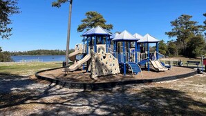 A playground with slides and climbing structures, surrounded by trees and a body of water in the background.
