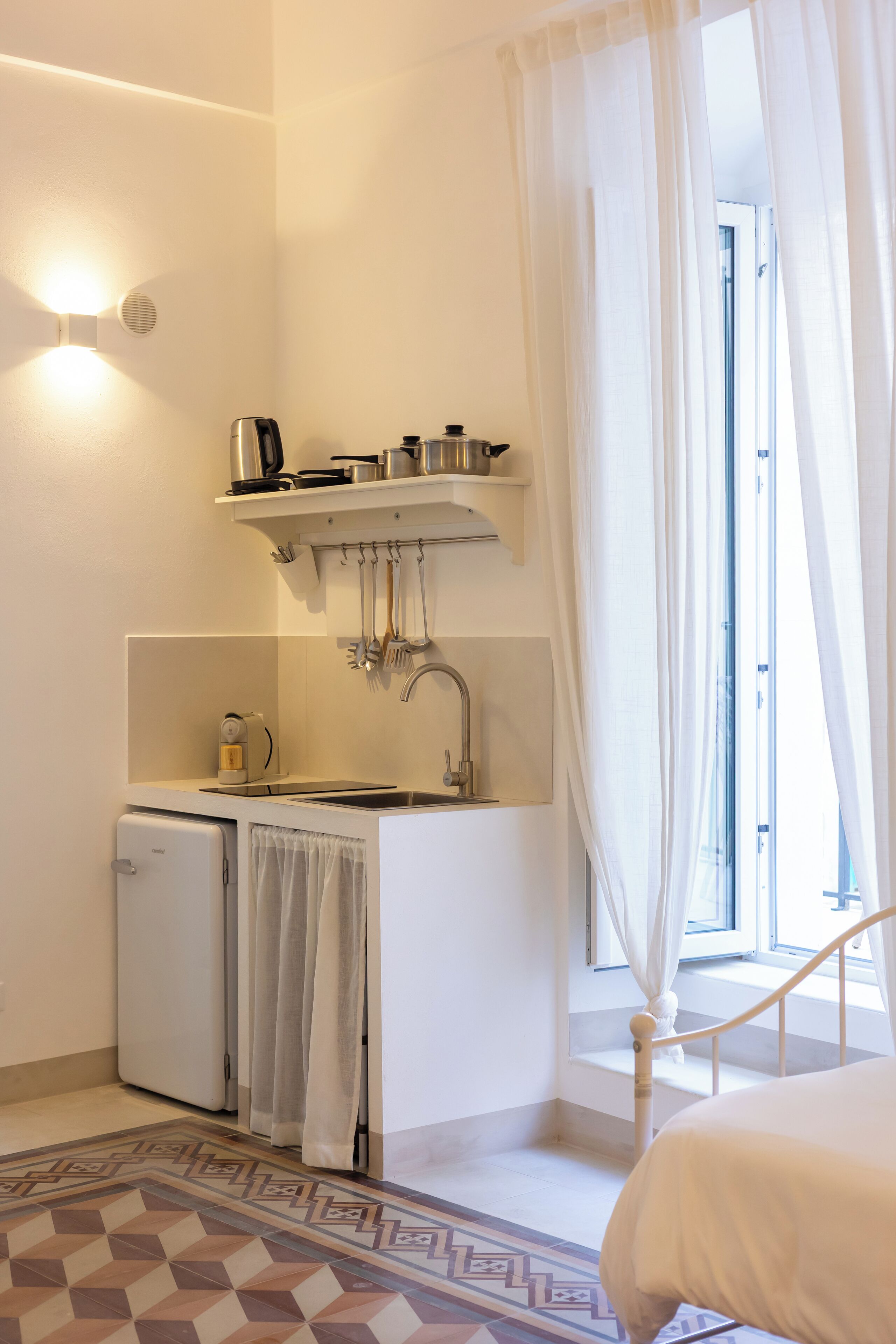 A compact kitchenette with a small fridge, a stove, and a sink. There is a window with sheer curtains and a bed with a white duvet in the foreground.
