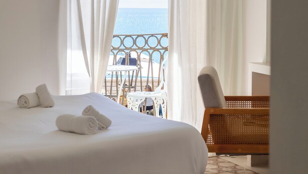 A hotel room with a bed, white curtains, a view of the sea, and a balcony with outdoor furniture.