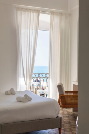 A hotel room with a bed, white curtains, a view of the sea, and a balcony with outdoor furniture. - Donna Veneranda (Vieste)