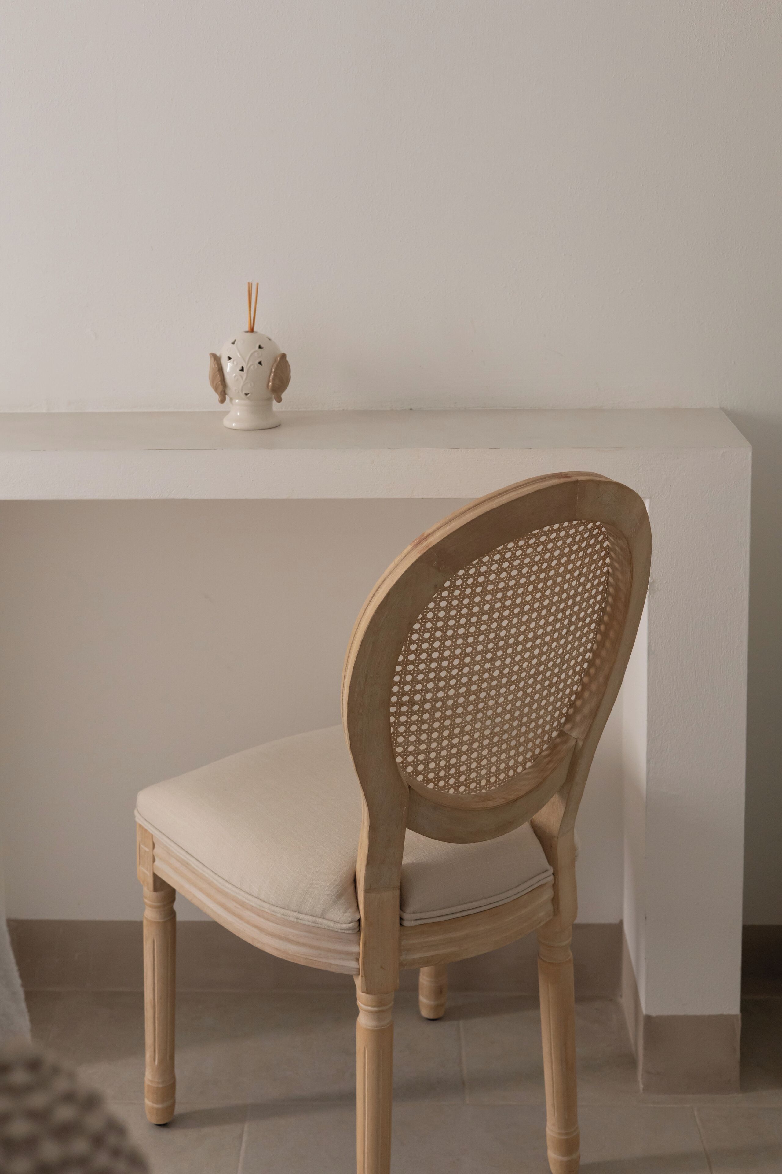 A wooden chair with a woven backrest and a cushioned seat, positioned in front of a white shelf with a small decorative item on top.
