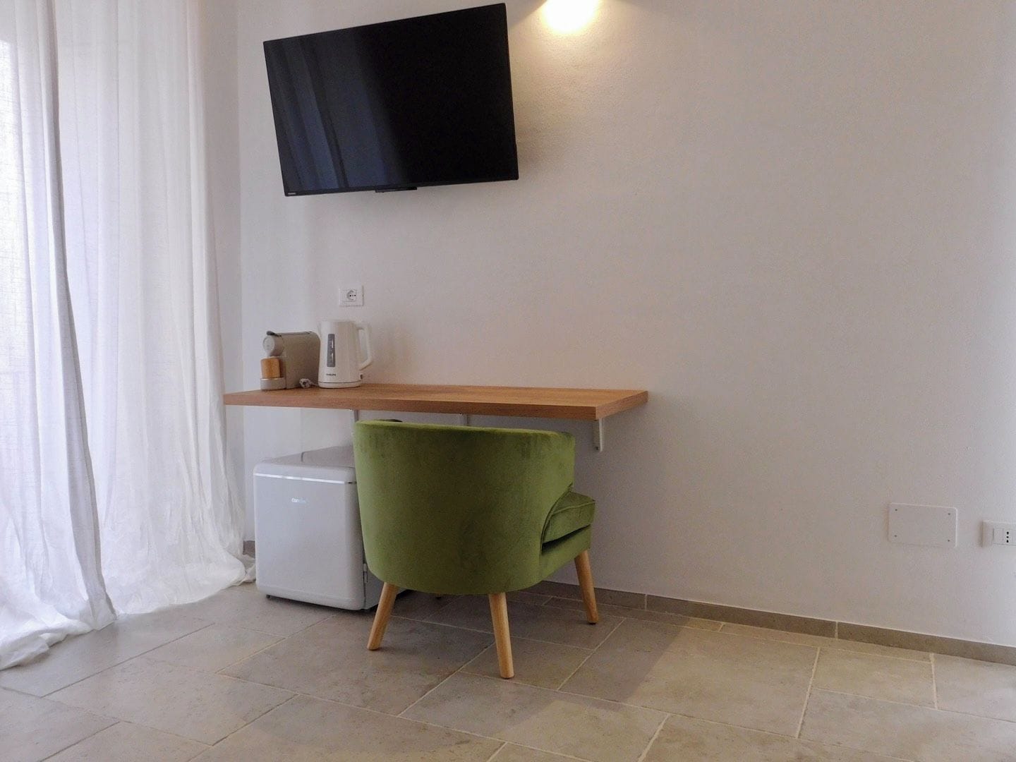 A small room with a flat-screen TV mounted on the wall, a wooden desk, a green chair, and a white mini-fridge.