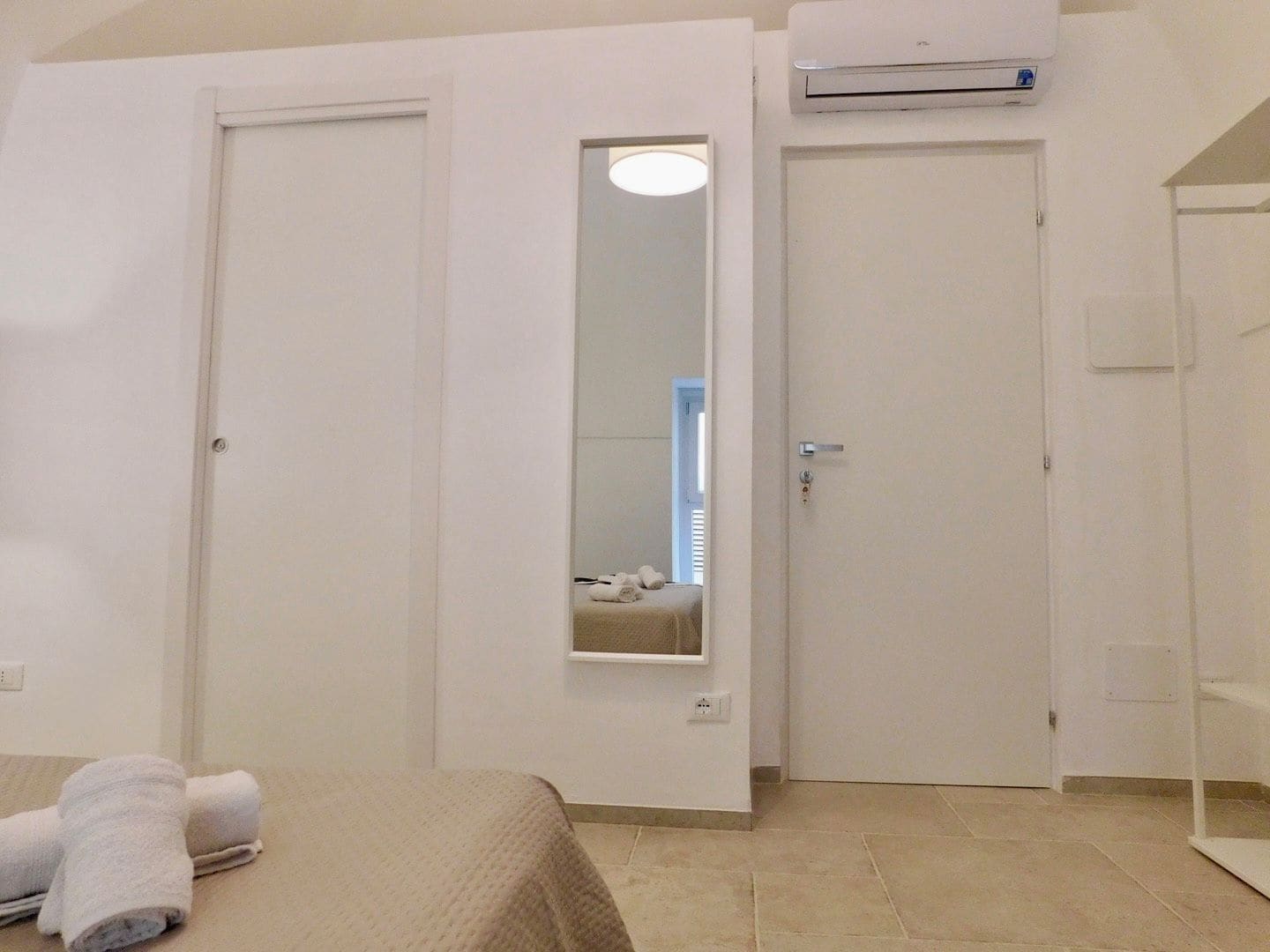 A hotel room with two doors, a bed with a beige bedspread, a wall-mounted air conditioning unit, and a small mirror.