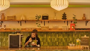 Ein Barista bereitet Getränke an der Theke zu, wo eine Kaffeemaschine, Regale mit verschiedenen Artikeln und ein Schild für Entwässerungsgetränke zu sehen sind.