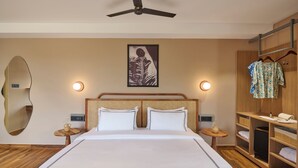 Ein Hotelzimmer mit einem Bett, einem Deckenventilator, einem an der Wand befestigten Spiegel, einem Nachttisch mit Lampe, einem gerahmten Bild an der Wand und einem Holzschrank mit Kleidung.