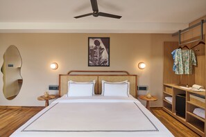 A hotel room with a bed, a ceiling fan, a wall-mounted mirror, a bedside table with a lamp, a framed picture on the wall, and a wooden wardrobe with hanging clothes. - Minimalist - The Varanasi Edit (Varanasi)