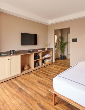 A modern hotel room with a wooden floor, a flat-screen TV mounted on the wall, a bed with a white headboard, and a small kitchenette area with a coffee maker and books. - Minimalist - The Varanasi Edit (Varanasi)