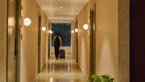 A hotel hallway with a person walking towards a window, a suitcase, and a plant in the foreground. - Minimalist - The Varanasi Edit (Varanasi)