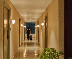 Un pasillo de hotel con una persona caminando hacia una ventana, una maleta y una planta al frente.