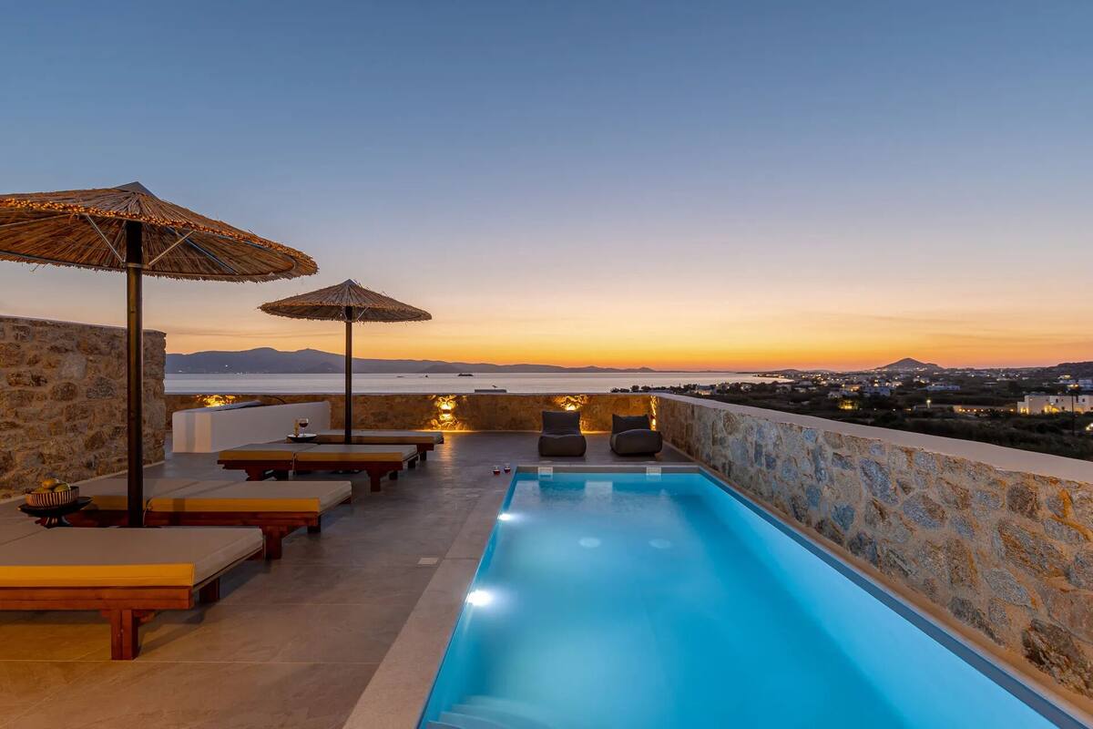 A rooftop pool with lounge chairs and umbrellas, overlooking a coastal cityscape at sunset.