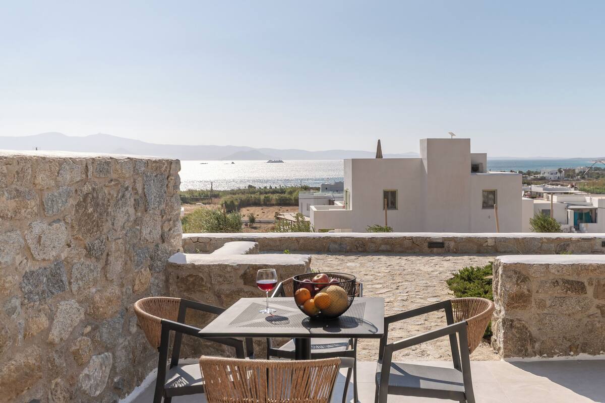 A terrace with stone walls, a table with a basket of fruit, a glass of red wine, and a view of the sea and distant mountains.