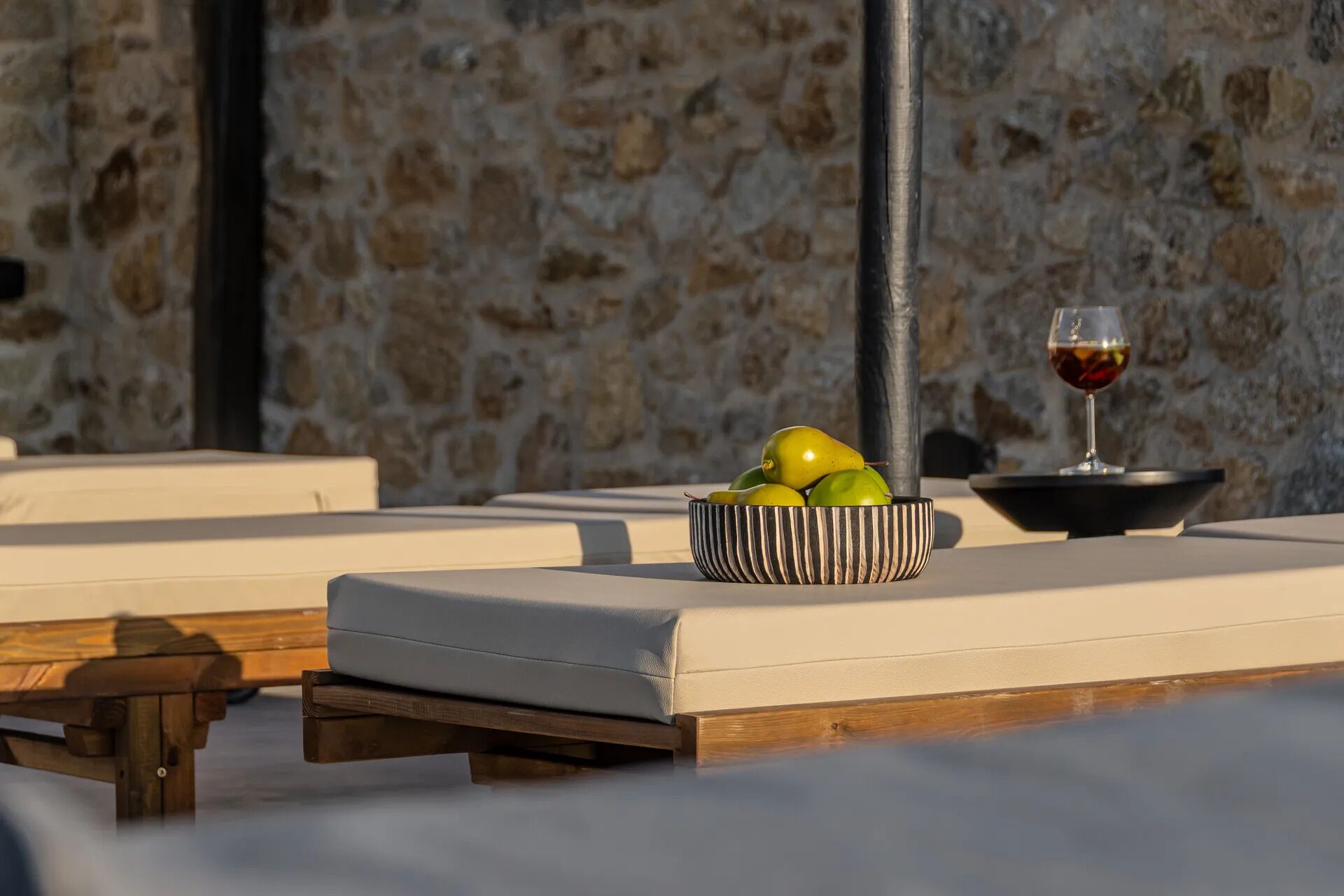 Un mur de pierres, un coin salon avec un saladier rayé rempli de fruits et un verre de vin rouge.