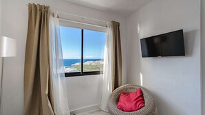 Een kamer met een groot raam, een rieten stoel met een rood kussen, en een flatscreen televisie aan de muur.