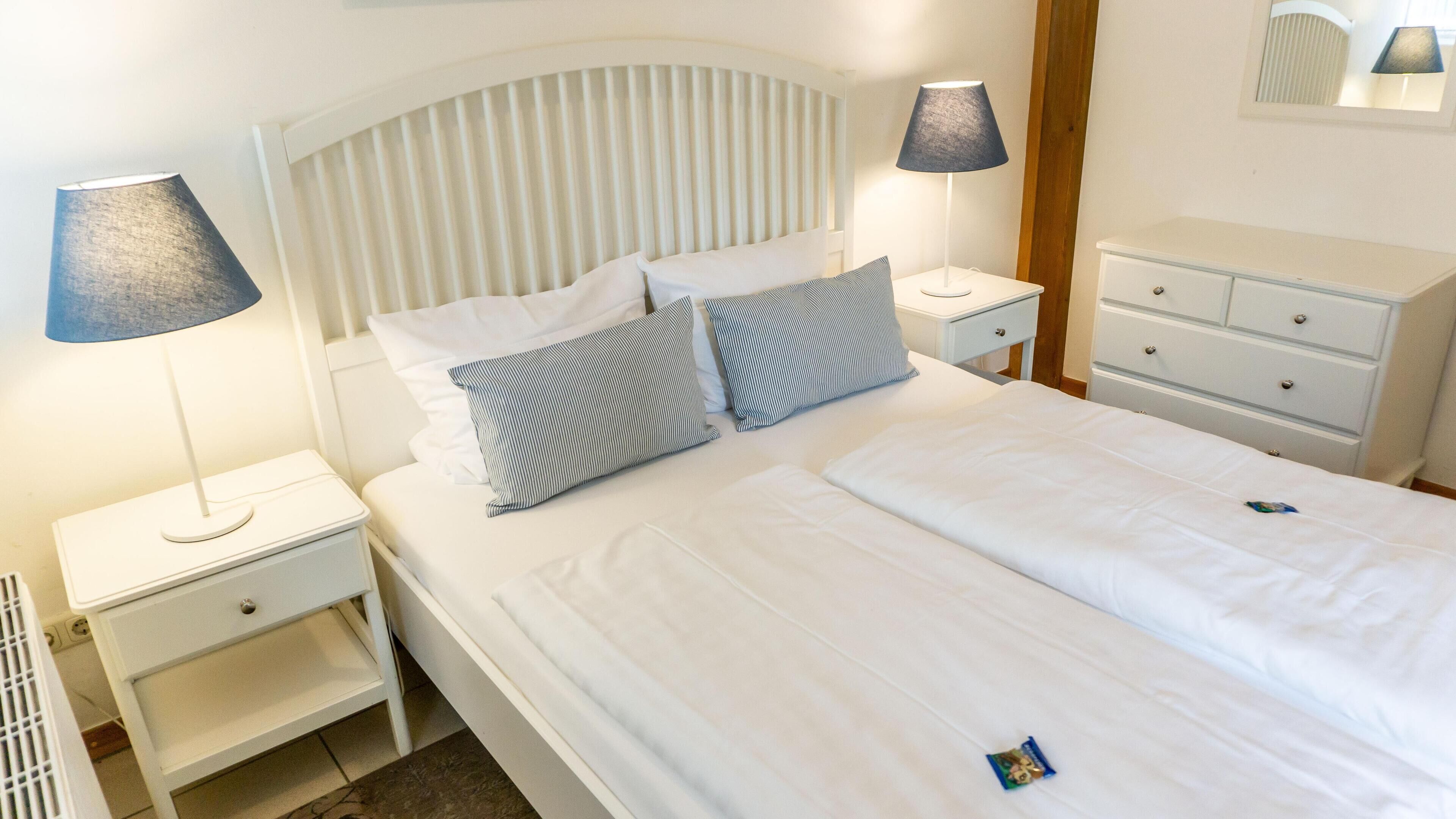 A neatly made bed with white bedding and striped pillows, a bedside table with a lamp, and a small drawer unit.