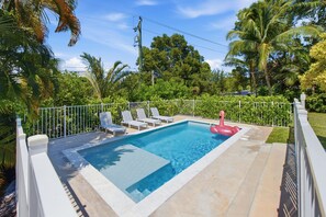 Piscina rectangular con una valla blanca, rodeada de palmeras y un cielo despejado.