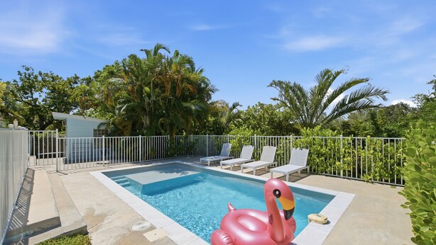 A swimming pool with a pink flamingo float, surrounded by a white fence and palm trees. - Welcome to Your West Palm Beach Getaway! (West Palm Beach)
