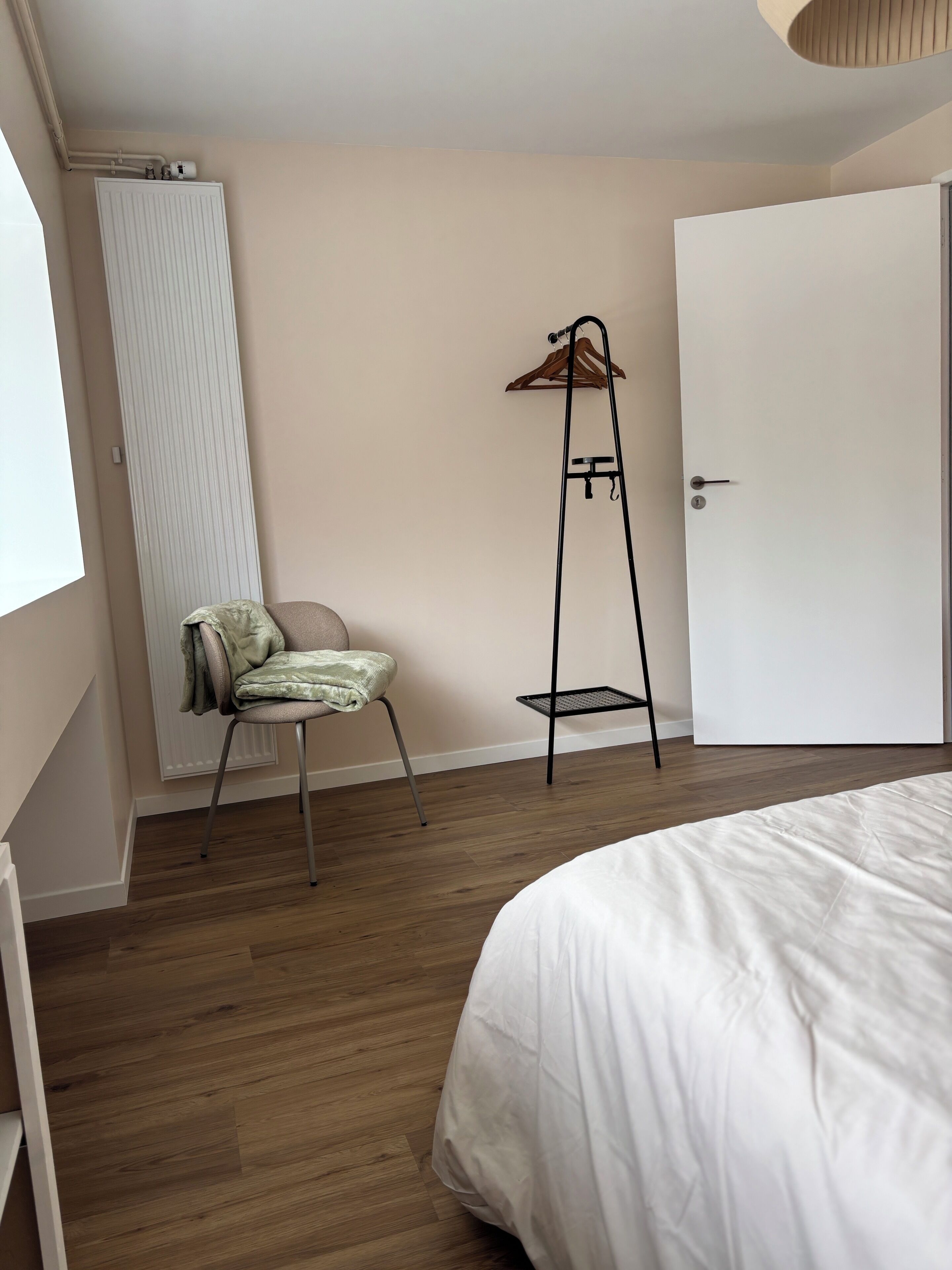 A bedroom with a bed, a chair, a radiator, a door, and a hanging rack.