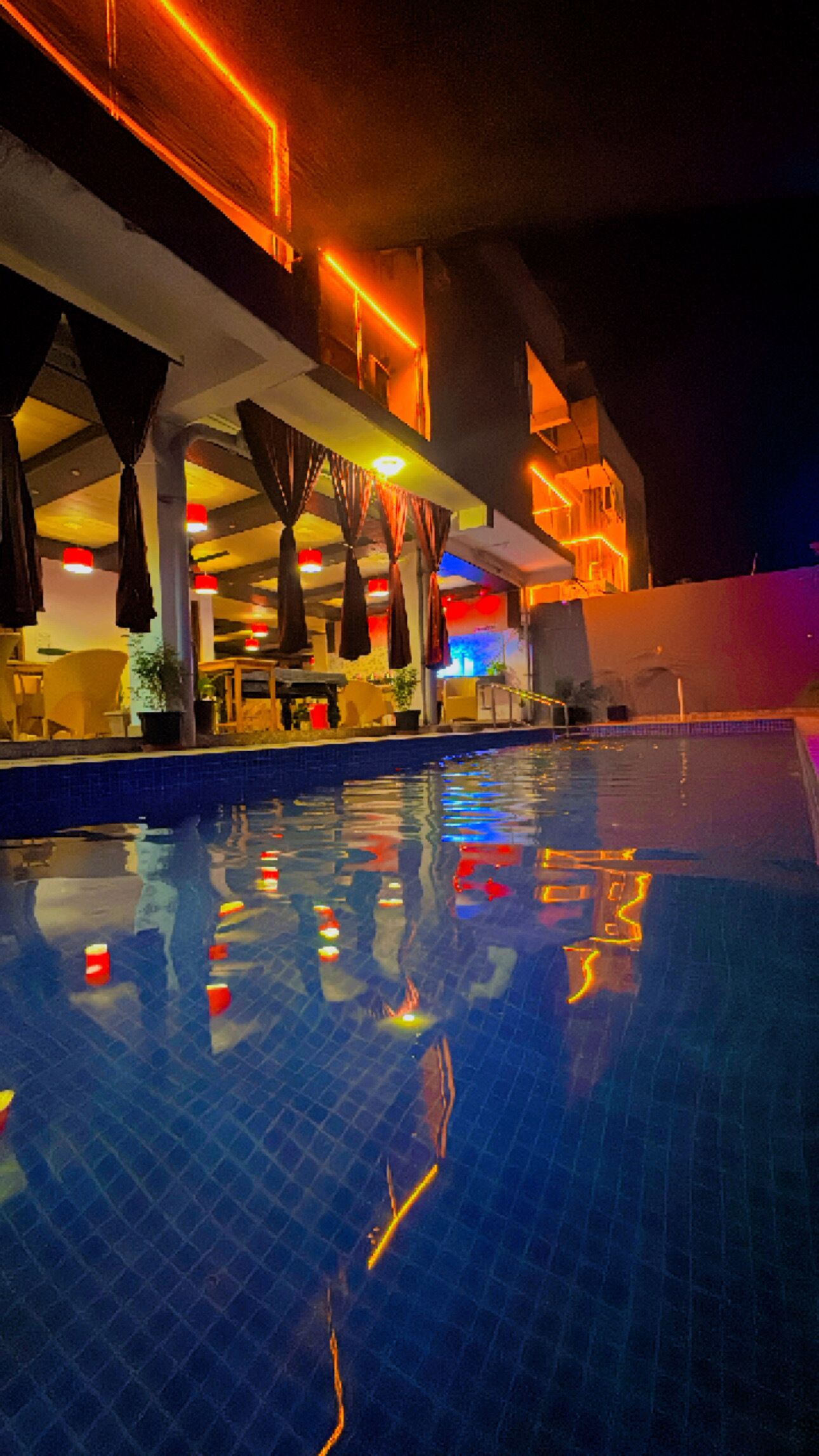 A nighttime pool area with a tiled floor, overhead lighting, and a covered seating area with umbrellas.
