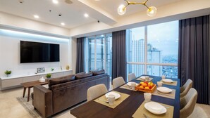 A modern living room with a dark brown sofa, a dining table set for four, and a large TV mounted on the wall. - Morich Suites Penthouse (Jakarta)