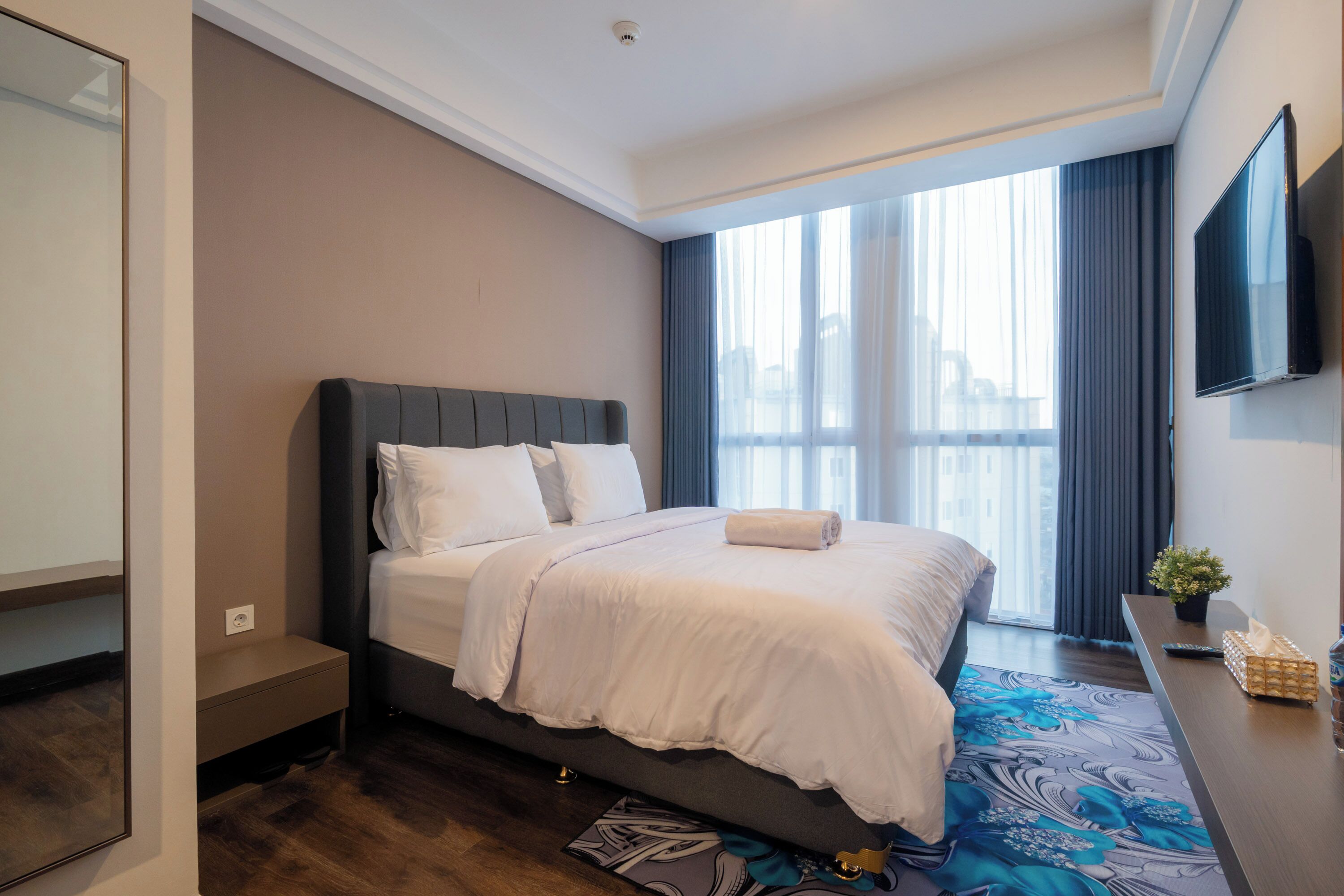 Una habitación de hotel moderna con una cama grande, un televisor de pantalla plana, una mesita con una planta y una ventana con cortinas.