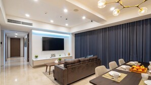 A modern living room with a dark brown sofa, a flat-screen TV, and a dining area with a table set for four. - Morich Suites Penthouse (Jakarta)