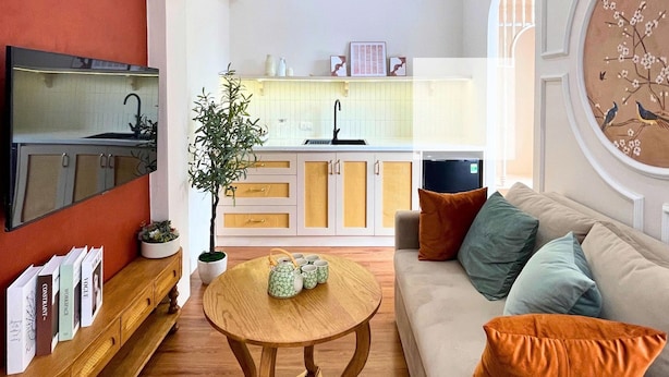 Ein Wohnzimmer mit einer Couch, einem Couchtisch und einem Fernseher. Im Hintergrund ist eine Küche mit Spüle und Schränken zu sehen.