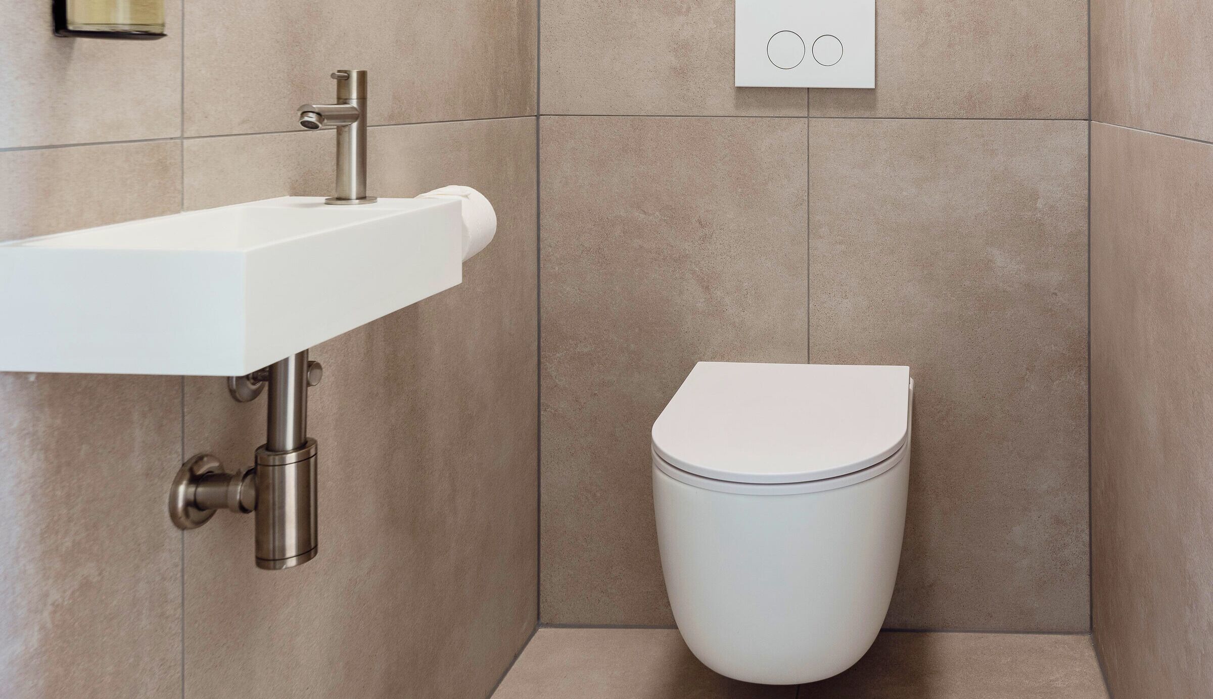 Ein Badezimmer mit weißem Waschbecken und WC, mit gefliesten Wänden und einem sichtbaren Rohr.