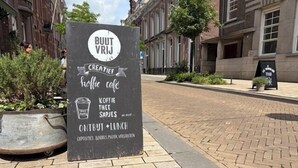 En skylt för ett kafé som heter ”BUUT VRIJ”, med en menytavla där kaffe, smörgåsar och lunchalternativ listas.
