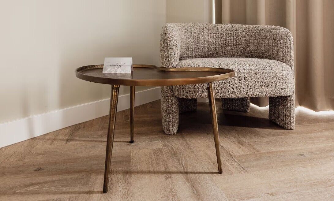 A round table with a wooden top and metal legs, placed next to a textured armchair in a room with wooden flooring and a curtain in the background.