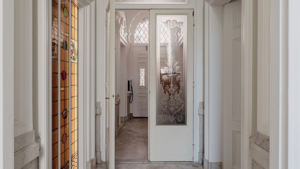 En hall med vita väggar, marmorgolv och utsmyckade dörröppningar som leder in till ett rum med en dekorativ glasdörr.