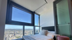 A bedroom with a large bed, a small table, and a chair, with a city view through large windows.