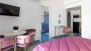 A modern hotel room with a pink bed, a flat-screen TV mounted on the wall, a small round table with a potted plant, and a pink chair. - Domus Vittorio double room with balcony and sea view  (Gaeta)