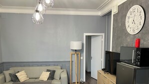Un espacio habitable compacto con sofá, una pequeña mesa, una lámpara, un reloj y una kitchenette con microondas y refrigerador.