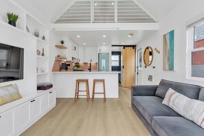 A modern kitchen with white cabinetry, a central island with stools, and a grey sofa in a bright living area. - Modern Beachfront Cottage (Oceanside)
