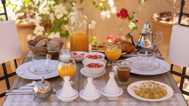 Une table de petit-déjeuner dressée avec divers plats, notamment du pain, des fruits et des boissons, dans un cadre ensoleillé en plein air.