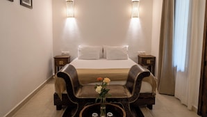 Une chambre d’hôtel comprenant un lit, deux tables de chevet, une petite table d’appoint, une chaise décorative, une petite table ornée d’un vase de fleurs et deux lampes fixées au mur.