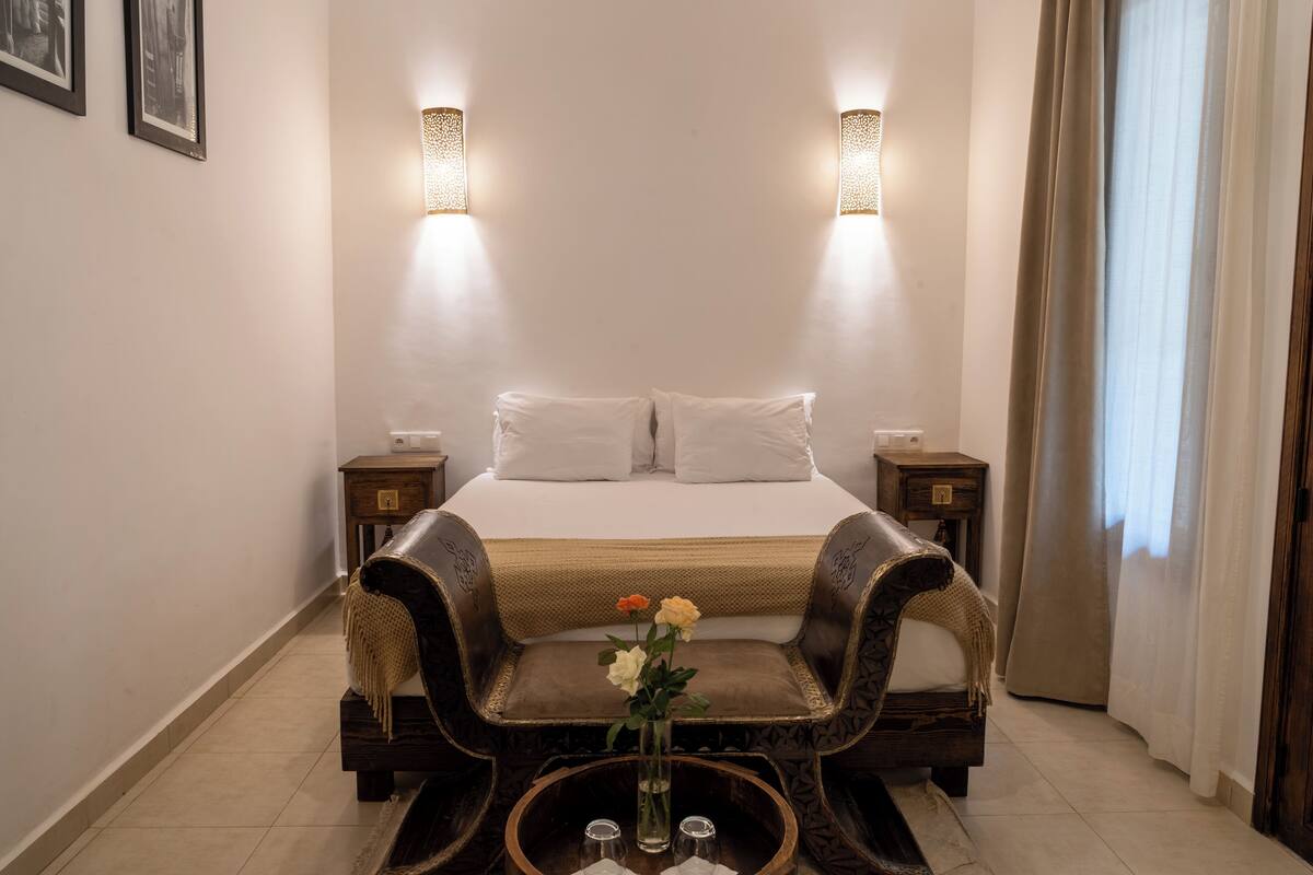 A hotel room with a bed, two bedside tables, a nightstand, a decorative chair, a small table with a vase of flowers, and two wall-mounted lamps.